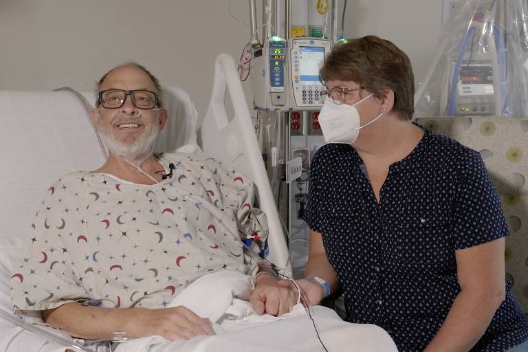 Homem de barba branca deitado em cama de hospital tem a mão segurada por uma mulher de máscara