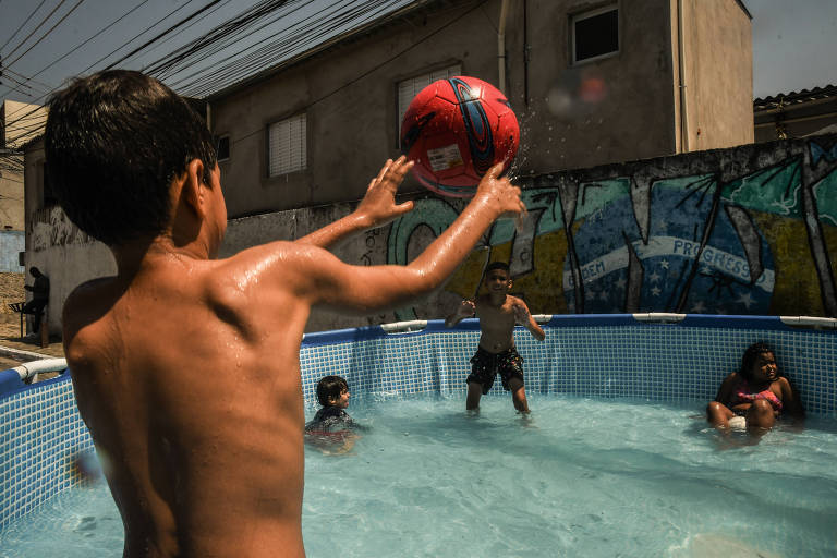 crianças jogam bola dentro de piscina de plástico armada em via pública; cenário urbano com muro, edificações e fios da rede elétrica ao redor.