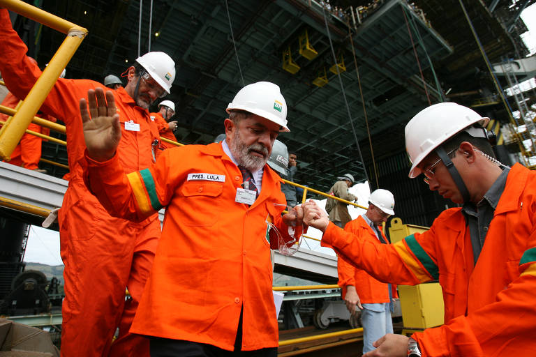 O presidente Lula, vestindo jaleco laranja e capacete branco cumprimenta outro homem com a mesma roupa, ambos com as mãos fechadas. Ao fundo aparecem estruturas metálicas e outros homens com os uniformes.
