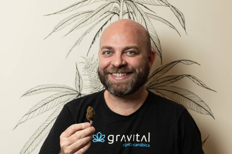 Retrato de Joaquim, um homem branco, careca, com barba curta; ele usa camiseta preta com o logo da empresa Gravital; sorrindo, ele segura um produto derivado de Cannabis; ao fundo, a parede tem o desenho de um pé de Cannabis sativa