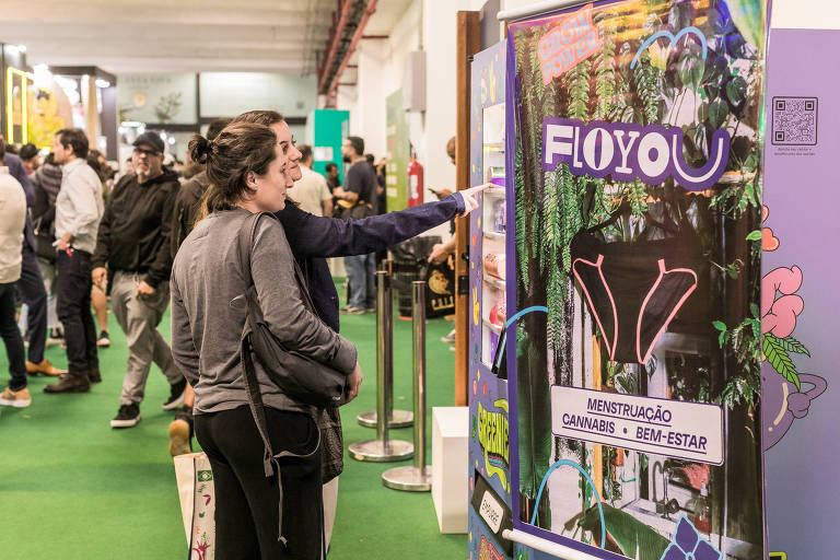 Mulheres apontam para máquina de produtos com pôster da marca FloYou, de calcinhas absorventes feitas de cânhamo, durante a ExpoCannabis Brasil
