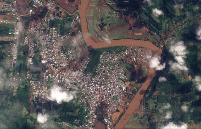 imagem de satélite mostra terreno depois de chuva, com lama nas ruas, transbordo do rio, que tem casas perto da margem 