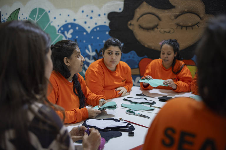 Foto mostra grupo de cinco mulheres à mesa, sob a qual se vê alguns materiais de costura. Quatro delas usa uniforme de cor laranja. Ao fundo, uma parede com grafite colorido