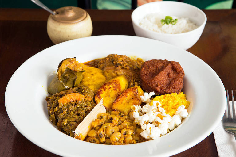 A foto mostra um prato branco com diversos itens: com caruru, vatapá, feijão fradinho, xinxim de galinha, acarajé, farofa de dendê, banana frita, pipoca e cana de açúcar. Ao fundo, uma pimenteira e uma tigela de arroz