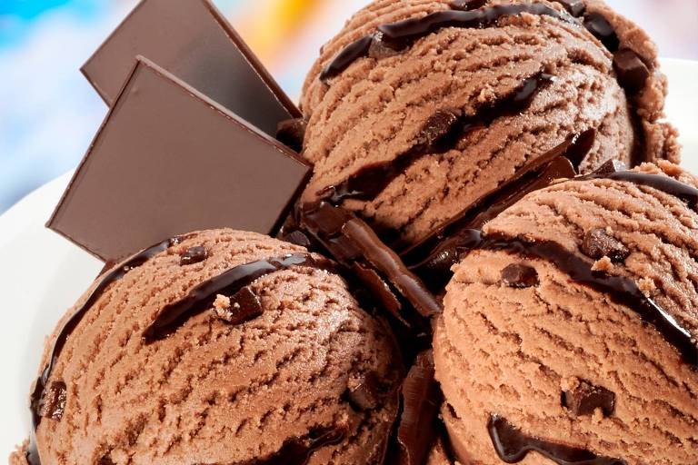 Imagem mostra três bolas de sorvete de chocolate, com calda do mesmo sabor, e dois tabletes de chocolate ao lado. O fundo da imagem é branco