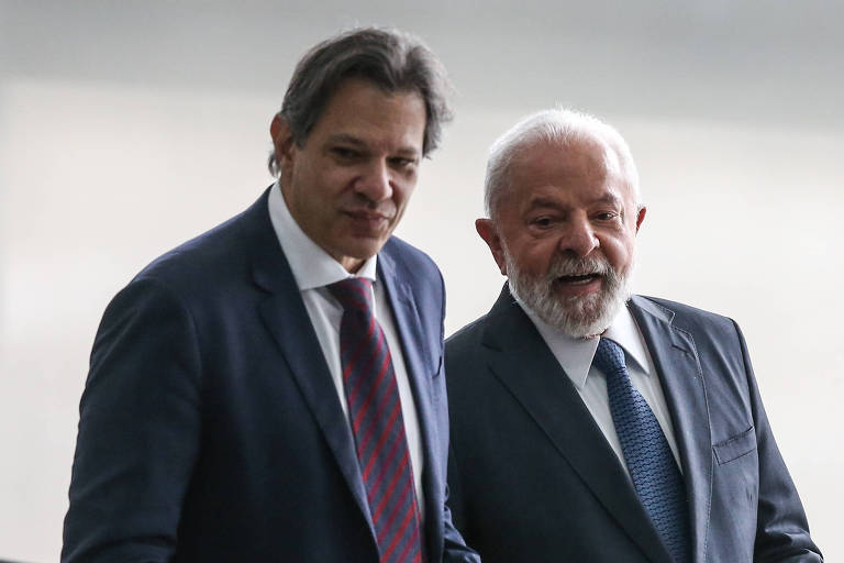 Haddad e Lula estão lado a lado em um ambiente formal. Haddad veste um terno escuro com gravata listrada, olhando para baixo com uma expressão séria. Lula, com barba branca e bigode, também veste um terno e olha para o mesmo ponto que o outro, exibindo uma expressão pensativa.