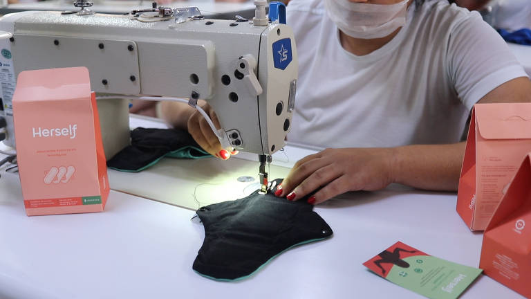 Foto foca em mulher trabalhando com máquina de costura. O rosco não é mostrado. Apenas o colo, tronco e braços. A mulher usa camiseta branca e máscara anti-covid
