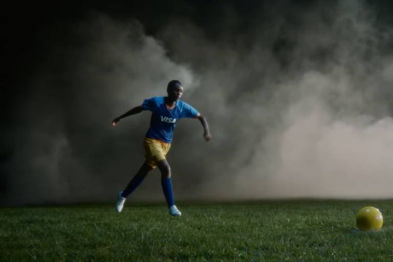 Jogadora de futebol negra se prepara para chutar a bola em estádio. Atrás dela, há uma névoa que cobre todo o fundo da imagem.