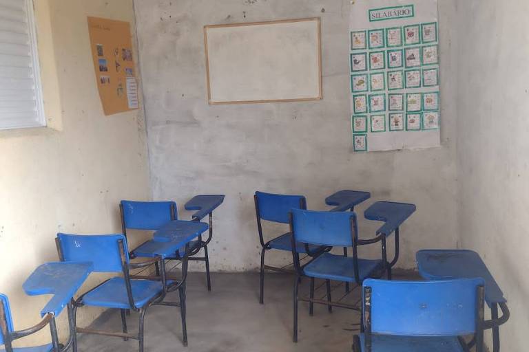 Pequena sala de aula improvisada, com algumas carteiras e cartazes na parede. Ao fundo, uma pqquena lousa