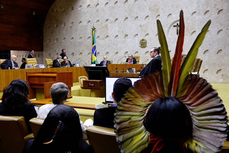 pessoa indígena de cocar está de costas, em primeiro plano; ao fundo, plenário do supremo tribunal federal, onde estão os ministros durante sessão