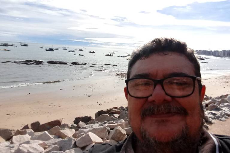 foto do tipo selfie mostra vanderli em primeiro plano. atrás dele está uma praia. ao fundo, o céu azul