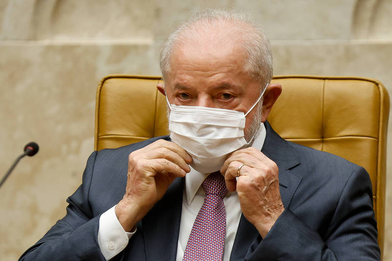 Lula está de terno e gravata e ajusta com as mãos máscara facial que cobre sua boca e nariz. Ele está sendo em cadeira do plenário do STF