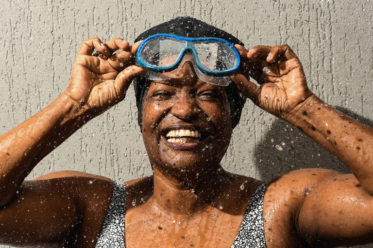 Fotografia colorida mostra um retrato focando o rosto de Luciana; ela é uma mulher negra e sorri para a foto; Luciana veste um maiô e segura um óculos de natação acima dos olhos; gotas de água caem sobre ela