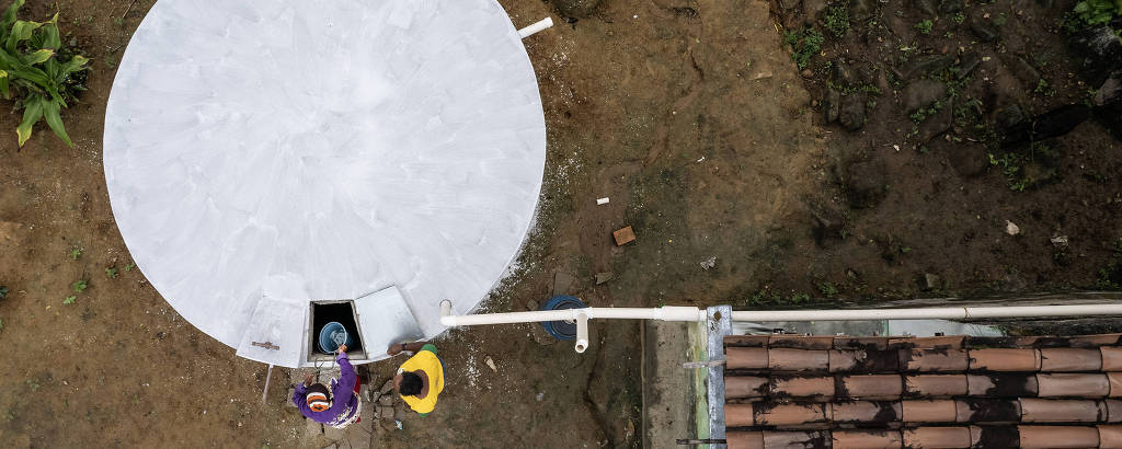 Imagem aérea de Cícero Claudino da Silva e sua mãe, Marinalva, pegando água cisterna recebida pela família: reservatório branco, redondo, de 16 mil litros