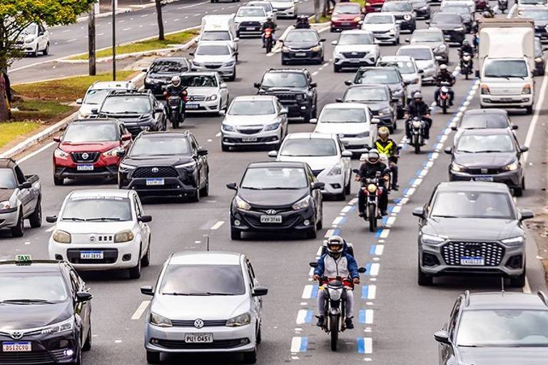 A imagem mostra um tráfego intenso em uma via urbana， com muitos carros e algumas motos. Os veículos estão em várias faixas， e a cena é capturada de uma perspectiva elevada. À esquerda， há uma área verde com árvores.