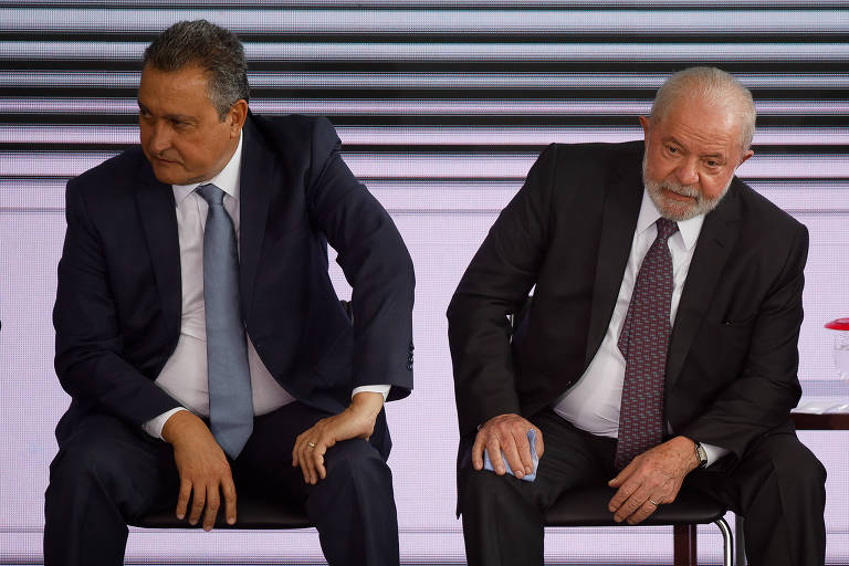 Rui Costa e Lula aparecem sentados, cada um se inclinando para um lado