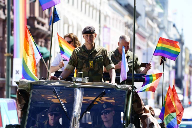 Homem com roupa militar em carro aberto ao lado de bandeiras arco íris