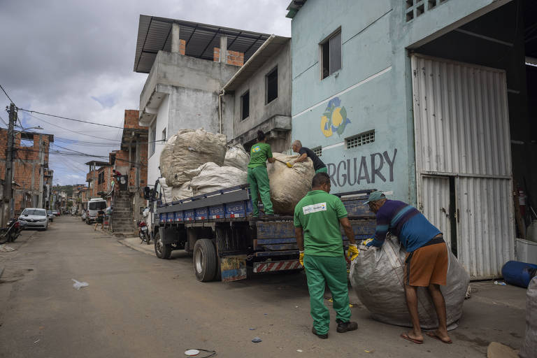 Foto em área externa mostra rua de comunidade periférica. Na imagem, o destaque é um caminhão com parte traseira cheia de sacos grandes de resíduos. Dois homens carregam mais um saco no veículo.