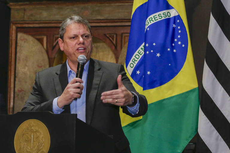 Tarcísio de Freitas discursa em evento de lançamento do programa Resolve Já. Cerimônia aconteceu no Palácio dos Bandeirantes, em São Paulo (SP). Tarcísio é um homem branco de cabelos grisalhos, estava vestido com um terno cinza escuro e camisa azul. Ele segurava um microfone na tribuna do auditório onde ocorreu o evento. Ao fundo, é possível ver uma parte da bandeira do Brasil e a parte preta e branca da bandeira do estado de São Paulo.