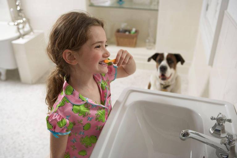 Menina escova os dentes na pia do banheiro, com um cachorro observando