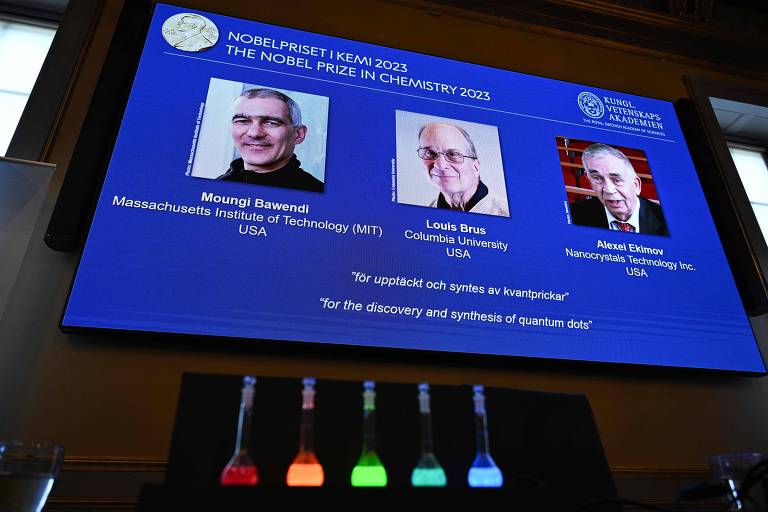 Os laureados com o Nobel de Química 2023 e, em frente à tela com seus rostos, frascos com líquidos brilhantes contendo a descoberta da qual participaram