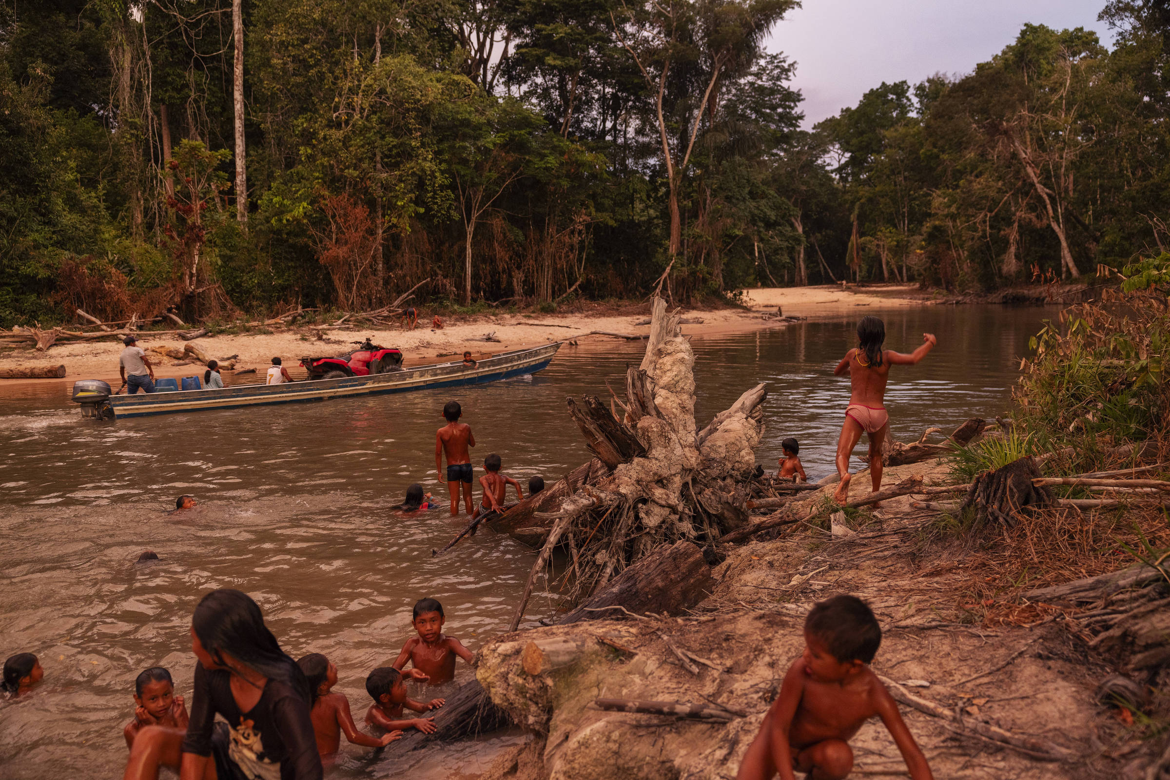 Crianças nadam na água e brincam na margem do rio enquanto uma embarcação passa mais ao fundo