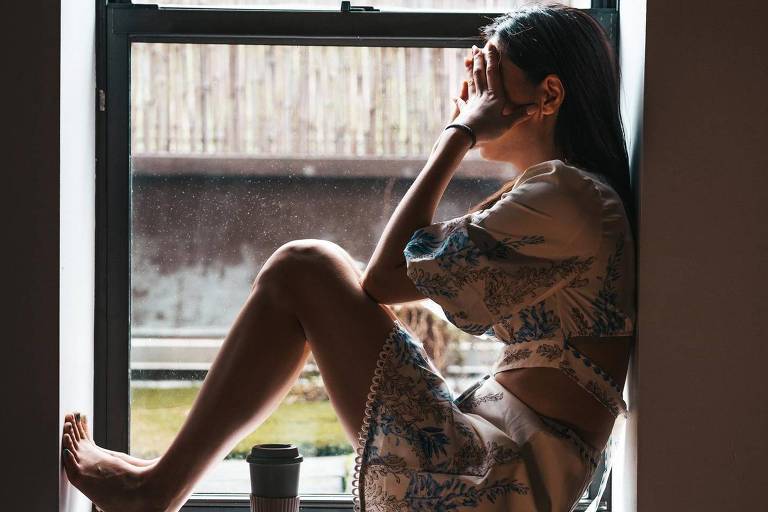 Uma mulher sozinha sentada em frente a uma janela com as mãos no rosto
