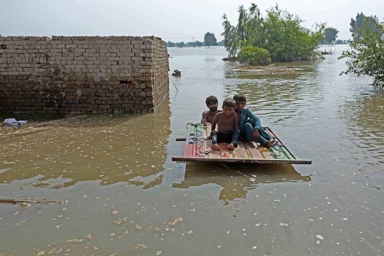 Duas crianças e um homem são evacuadas, sobre uma placa de madeira que flutua na água, durante enchente no Paquistão