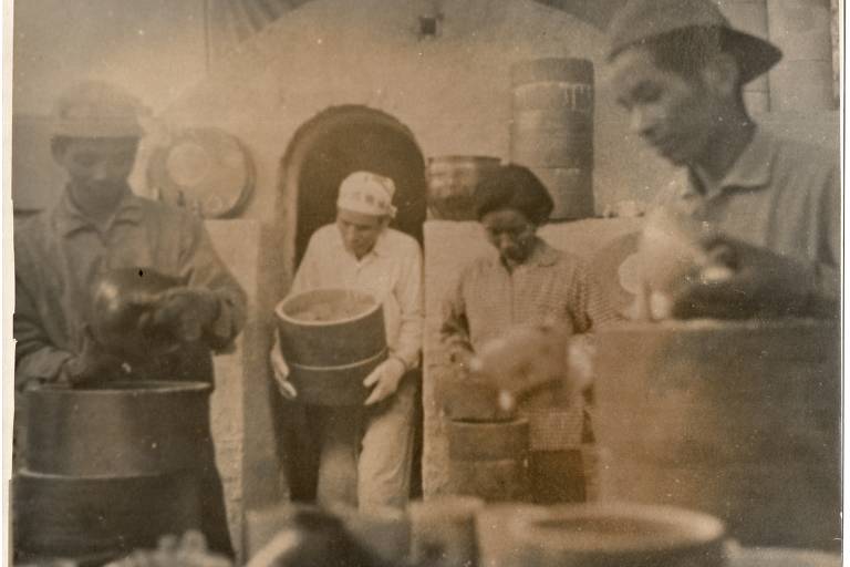Um homem japonês com luvas e lenço sobre a cabeça retira uma caixa arredondada do forno. Uma mulher e dois jovens japoneses estão perto, olhando para outros produtos retirados do forno.