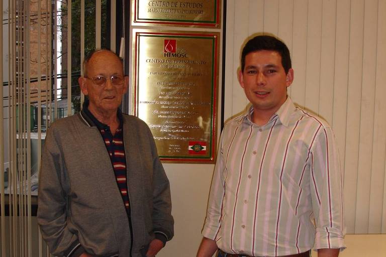 O médico Mário Roberto Kazniakowski, ao lado de seu filho, Mário Roberto Júnior, no Hemocentro de Florianópolis, com uma placa no fundo 