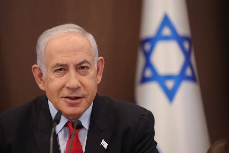 Benjamin Netanyahu fala ao microfone; há uma bandeira de Israel desfocada no fundo