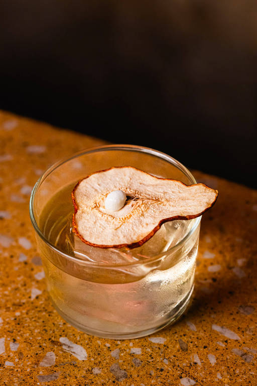 A foto mostra um copo baixo com drinque feito com tequila, purê de pera e vem decorado com uma fatia da fruta desidratada sobre um cubo grande de gelo