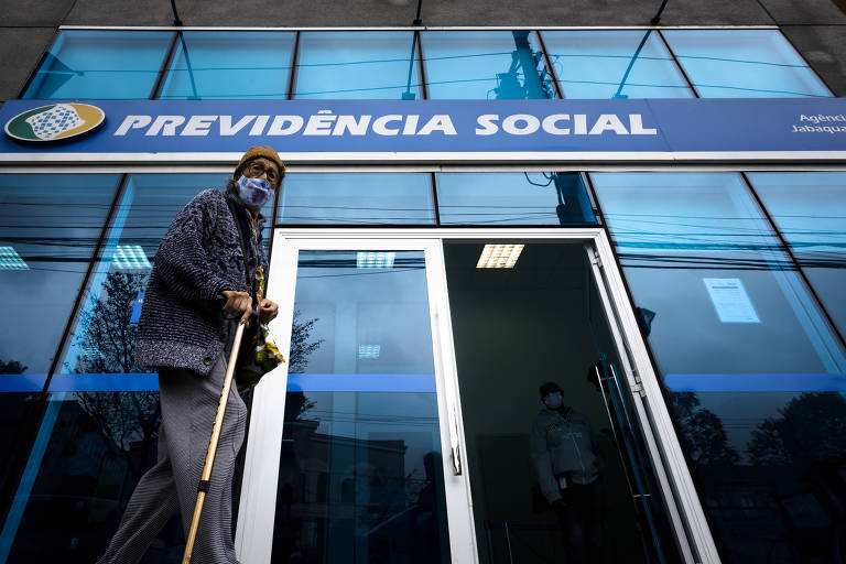 Idoso com máscara, gorro e bengala caminha em frente a prédio envidraçado azul, com logotipo da Previdência Social