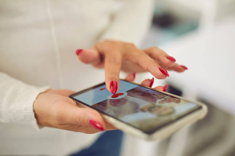 Uma mão feminina com unhas vermelhas está tocando a tela de um smartphone. A tela exibe imagens de duas pessoas. A mão está posicionada sobre o dispositivo， que parece estar em um ambiente interno com fundo desfocado.