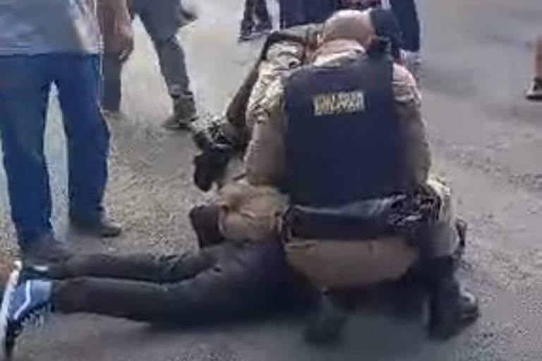 A foto mostra um policial militar ajoelhado tendo à sua frente, deitado, imobilizado, autor de ataque a facadas na saída de escola em Poços de Caldas.