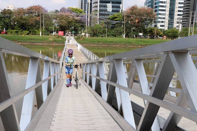 passarela sobre o rio, foto é feita de uma margem e mostra a outra ao fundo, ciclista caminha levando bicicleta 