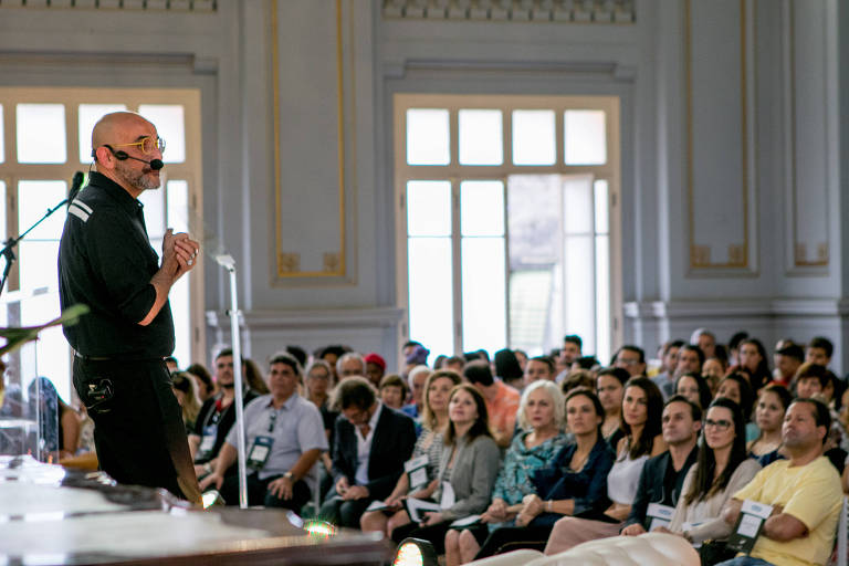 Em evento corporativo, homem fala à plateia com microfone. Foto mostra palestrante em palco com roupas pretas e, à sua frente, uma plateia com cerca de cem pessoas, sentadas em cadeiras.