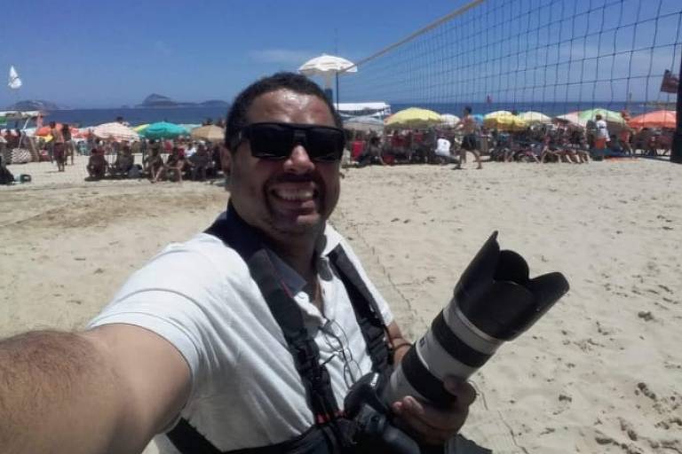 robson faz selfie, está de óculos escuros, camiseta branca, segura máquina fotográfica. ao fundo, areia e uma rede de futevôlei