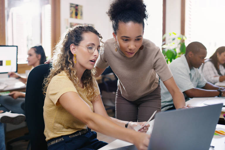 Fotografia colorida mostra um escritório com pessoas sentadas trabalhando em computadores; em descaque uma mulher branca de cabelos cacheados mostra algo na tela de um computador para uma mulher negra, que observa de pé