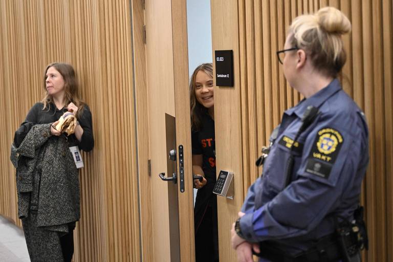Greta espia sorrindo abrindo uma porta; do lado de há duas mulheres