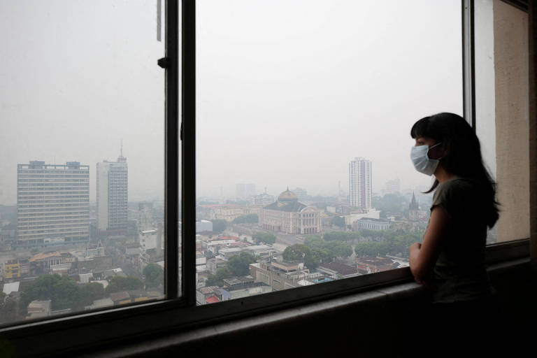 Mulher usando máscara dentro de apartamento observa vista cinzenta de cidade enfumaçada