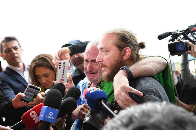 Homem loiro e de barba ao lado do pai, que chora; ambos estão cercados de jornalistas e microfones da imprensa