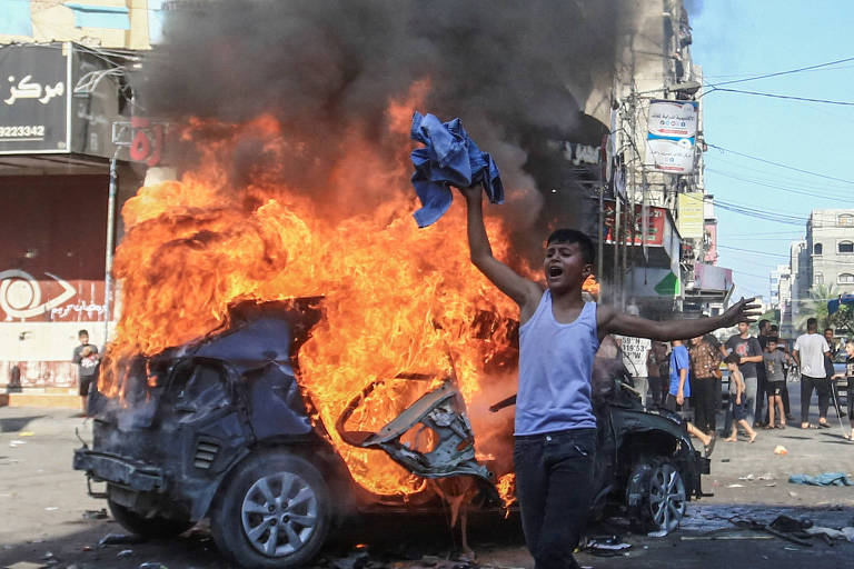 menino agita pano e grita , ele esta à frente de um veículo que está pegando fogo, atrás dele há pessoas e prédios