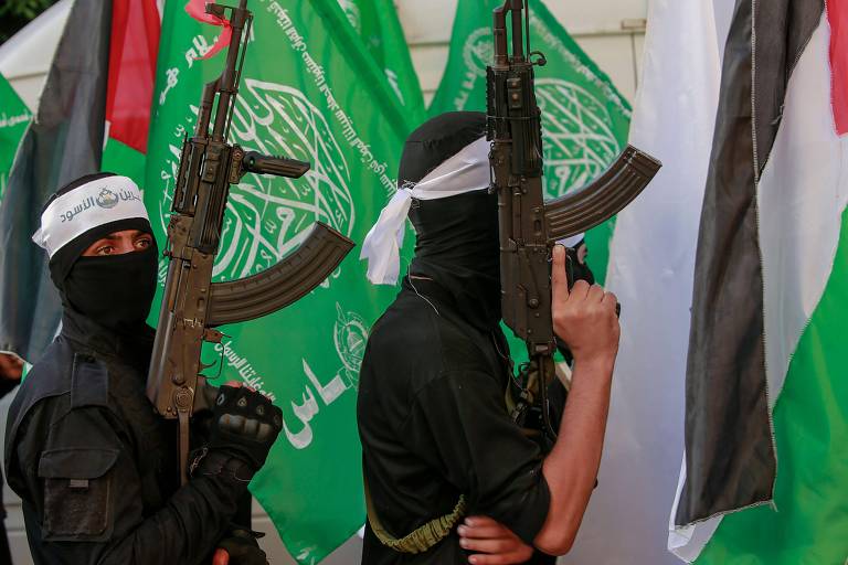 homens vestidos com farda militar usam balaclava e empunham fuzis. atrás deles estão as bandeiras da palestina e do hamas