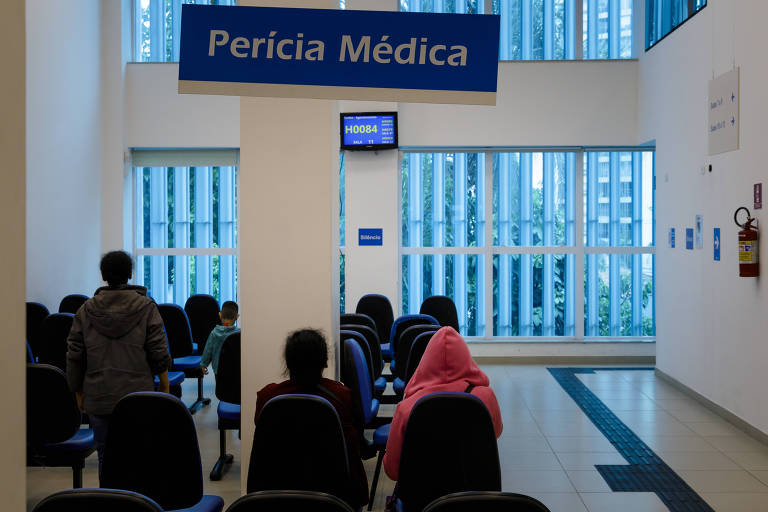 Interior de agência do INSS mostra pessoas sentadas em cadeiras azuis; há uma placa azul em que está escrito "Perícia Médica" em branco. 