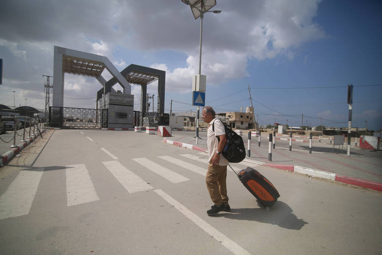 homem caminha puxando mala de viagem que tem rodinhas por uma rua. ao fundo, a passagem de Rafah, que tem duas estruturas em formato de arco por cima dos portões. 