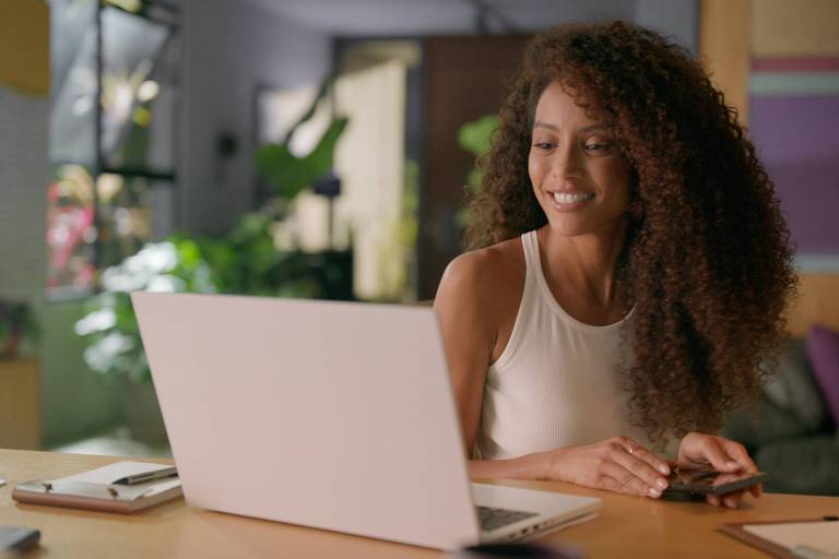 Taís Araújo está sentada sorrindo de frente para um laptop. Seus cabelos cacheados estão soltos e ela usa uma blusa de manga curta