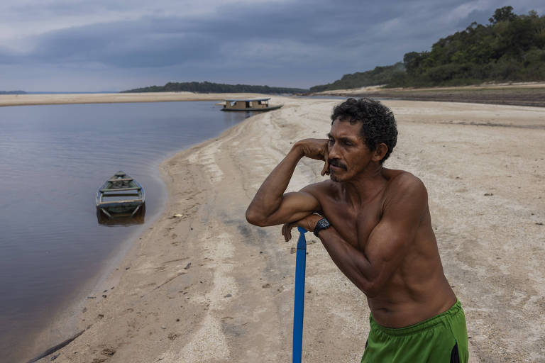 Homem apoiado em cabo de vassoura em uma praia do rio; ao fundo, há uma canoa