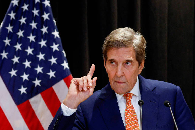 Kerry aponta com o indicador da mão direita ao lado de uma bandeira dos EUA