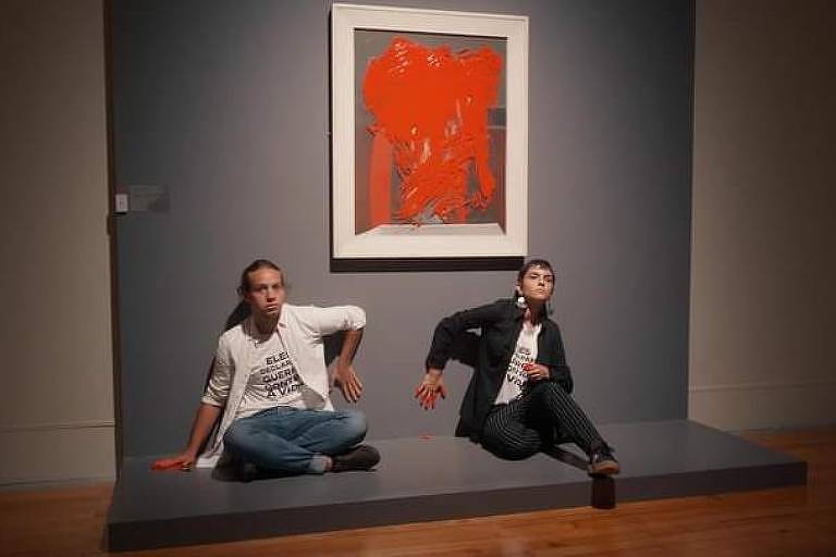 Dois jovens posam com uma mão na parede, sentados no chão, com o quadro preso à parede manchado de tinta vermelha
